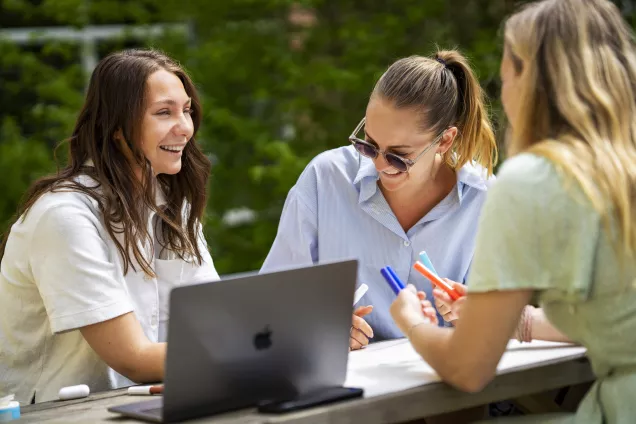Tre unga kvinnor sitter utomhus vid ett bord med en uppfälld laptop och studerar skrattande. Foto