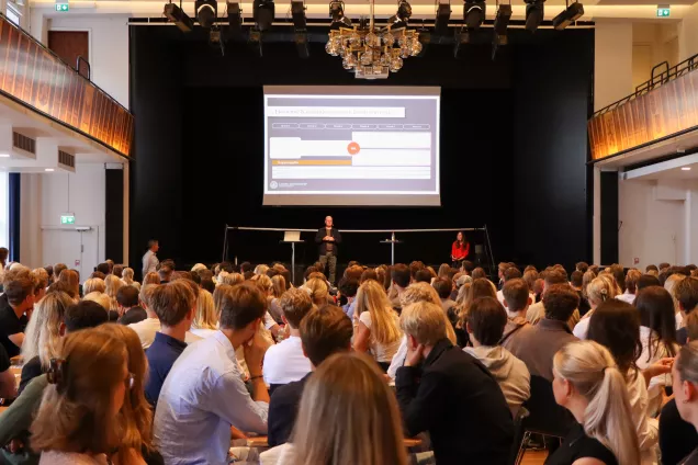 Vacker gammal aula full av studenter, lärare på scen presenterar. Foto. 