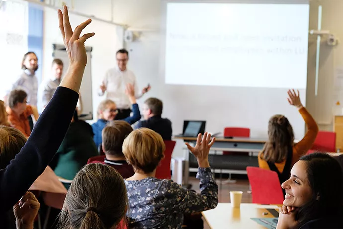 Klassrum och personer bakifrån med uppsträckt hand i förgrunden. Foto